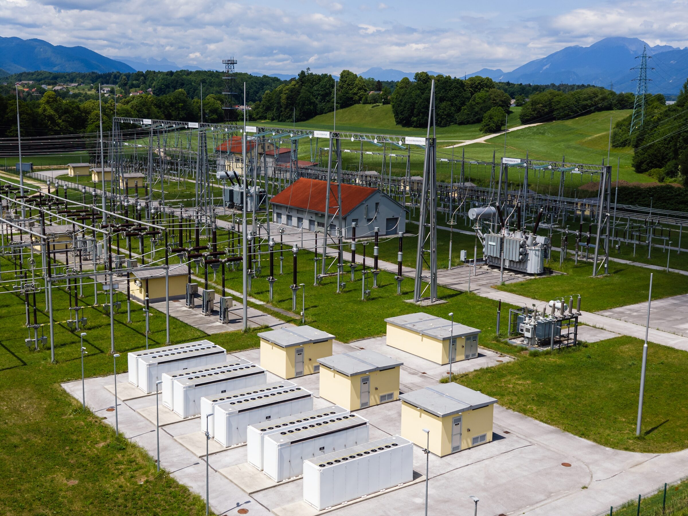 Battery containers on a concrete pad inside a substation, with green hills and mountains in the distance